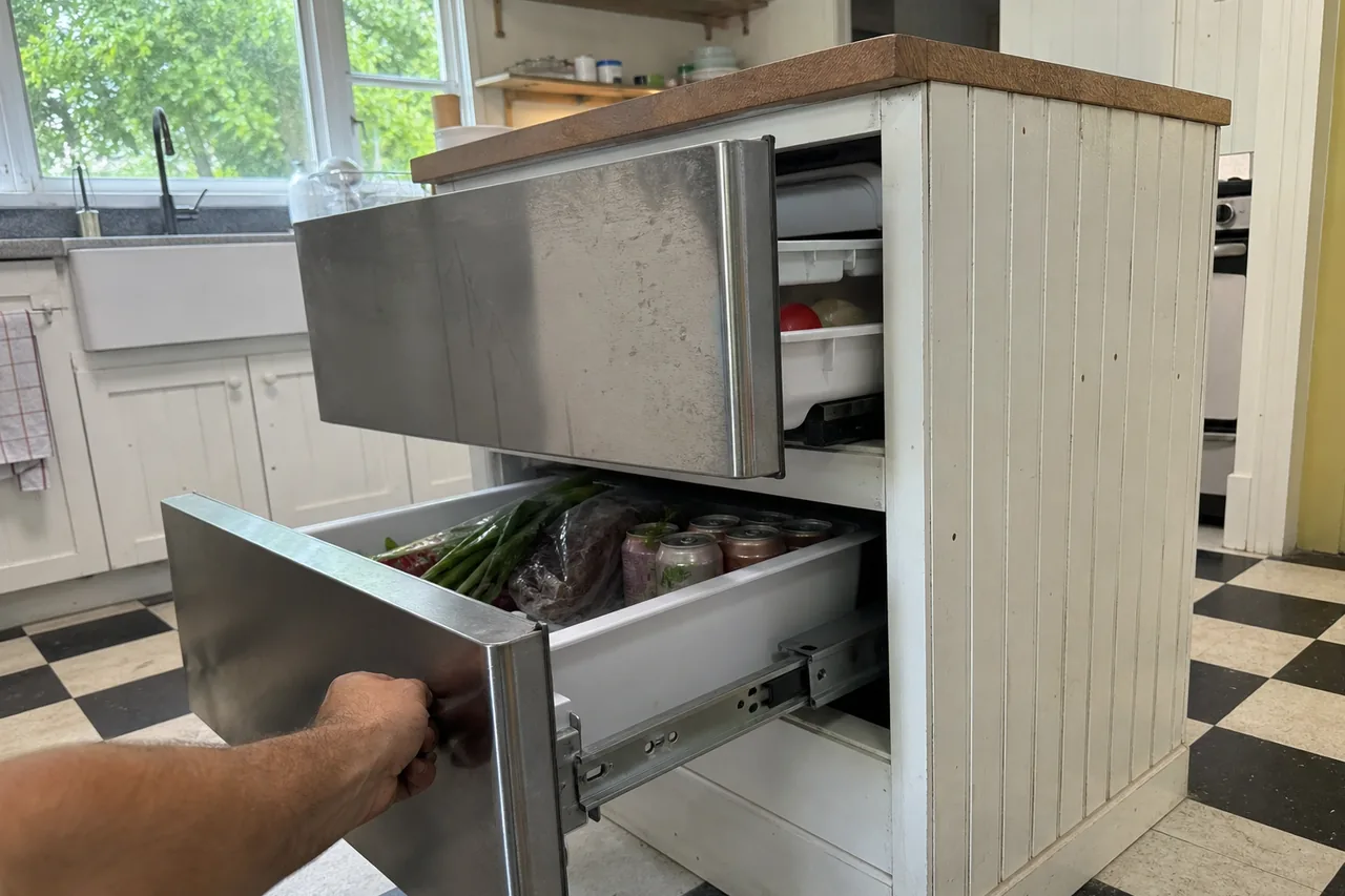 Two stainless refrigerator drawers set into a white beadboard island, the lower one pulled out on its steel runner to show scallions, a bag of greens and cans.