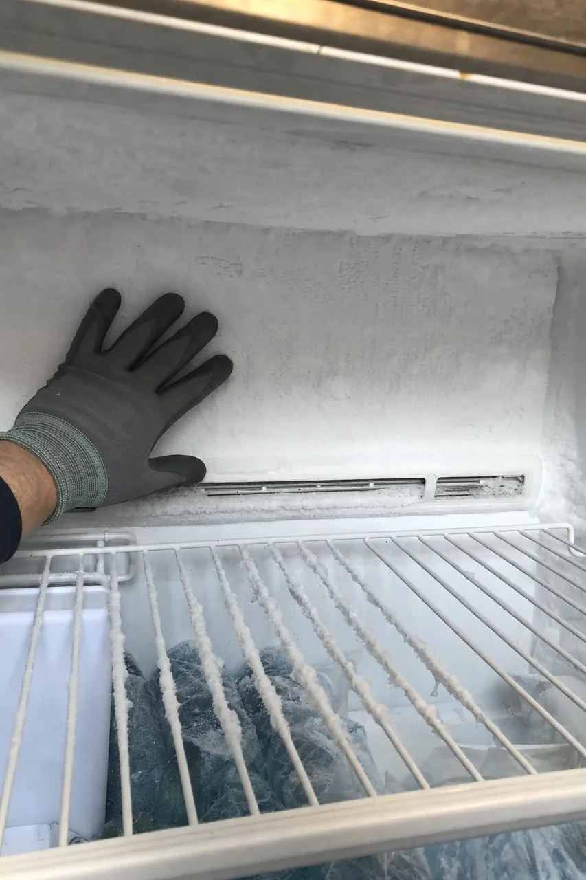 The inside rear wall of a freezer compartment sheeted in white frost, a gloved hand resting against it and the vent grille half buried in ice.